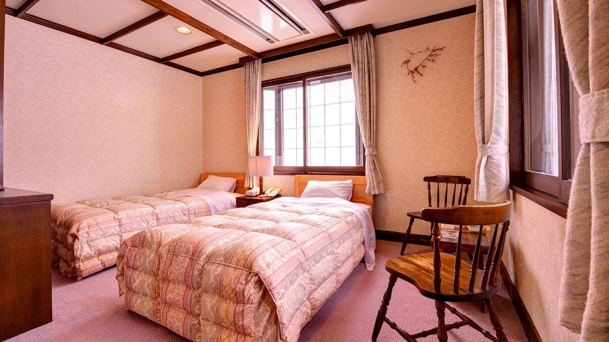 A hotel room with two beds, a wooden chair, a nightstand, and a window with curtains.