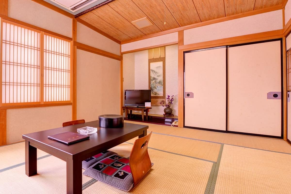 Ein traditioneller japanischer Raum mit Tatami-Boden, einem niedrigen Tisch mit einem Laptop, einem Fernseher und Schiebetüren.