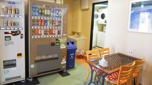 Un distributeur automatique de boissons et d’en-cas, une poubelle, une table avec des chaises et, au fond, une machine à laver.