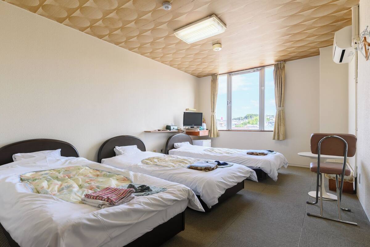 A hotel room with three beds, a desk with a computer, a chair, and a window with a view.