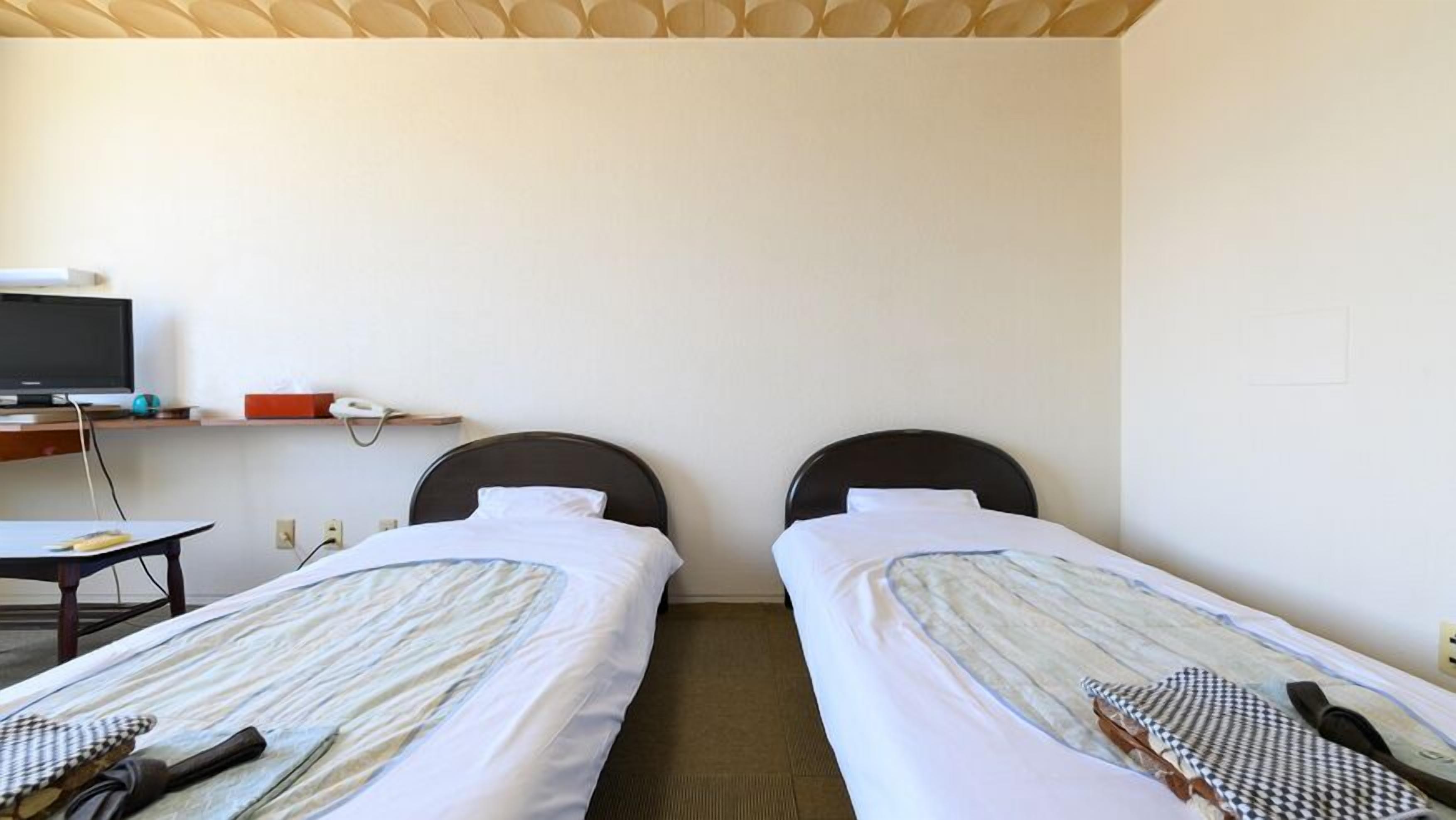 Quarto de hotel com duas camas de solteiro, uma escrivaninha com computador e telefone, e uma televisão sobre um suporte.