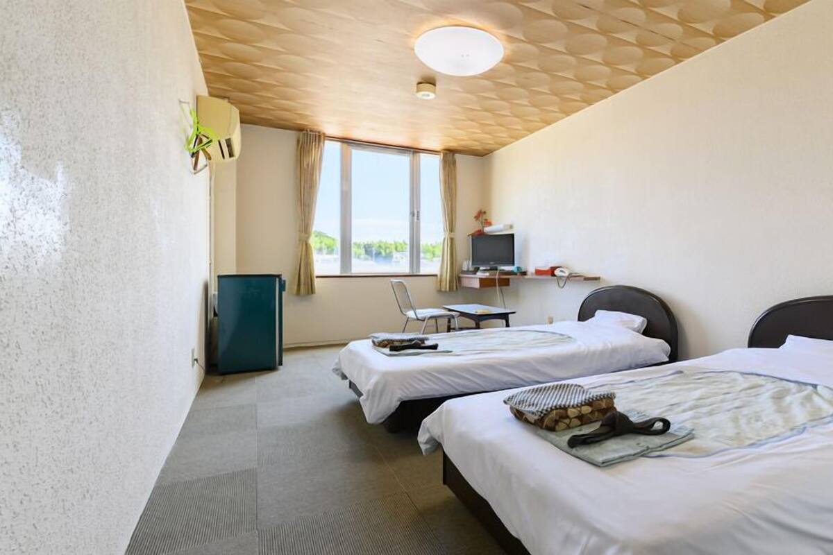A hotel room with two beds, a desk with a computer, a small fridge, and a window with curtains.