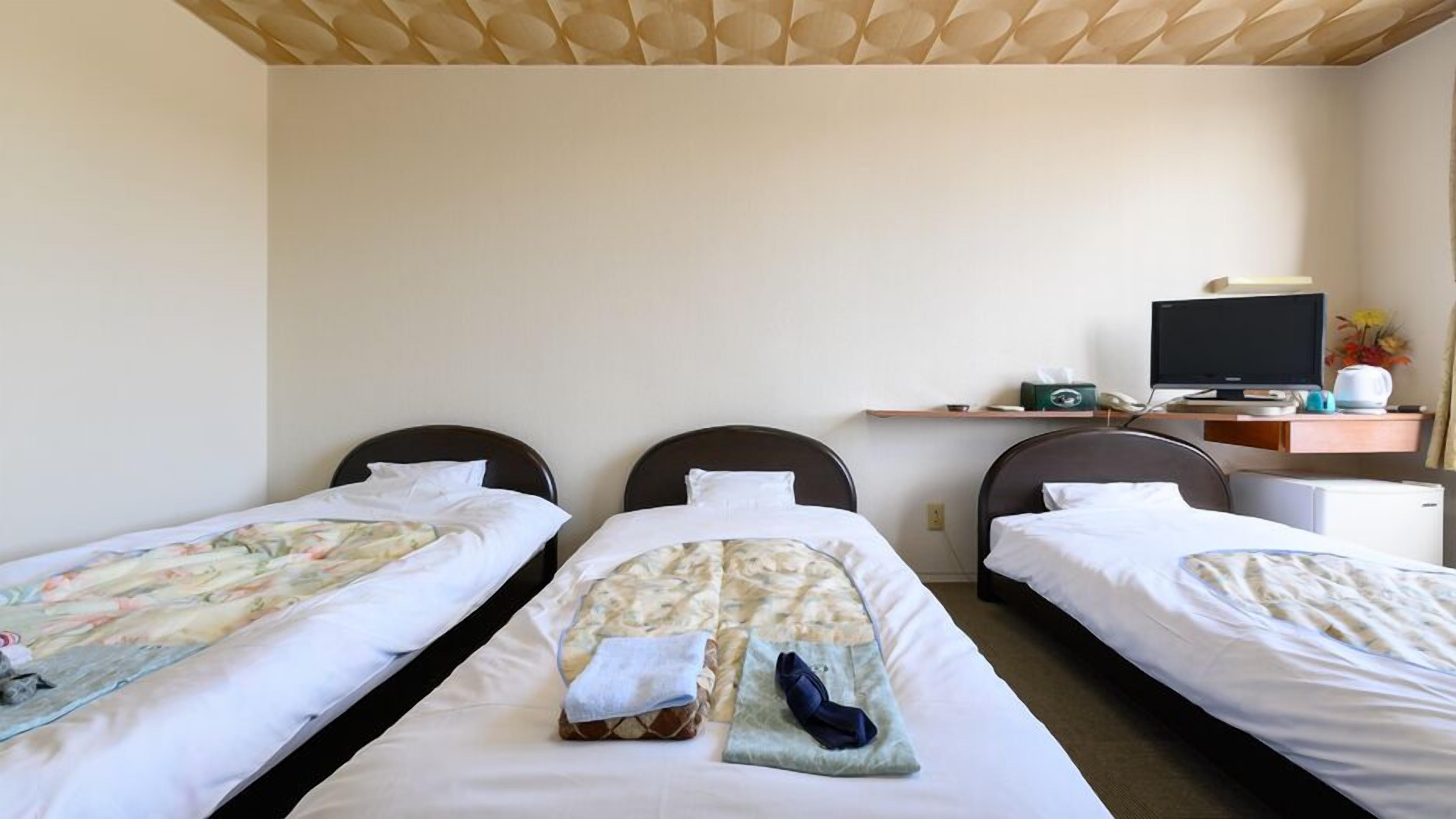 Quarto de hotel com três camas de solteiro, uma escrivaninha com computador e uma pequena geladeira.