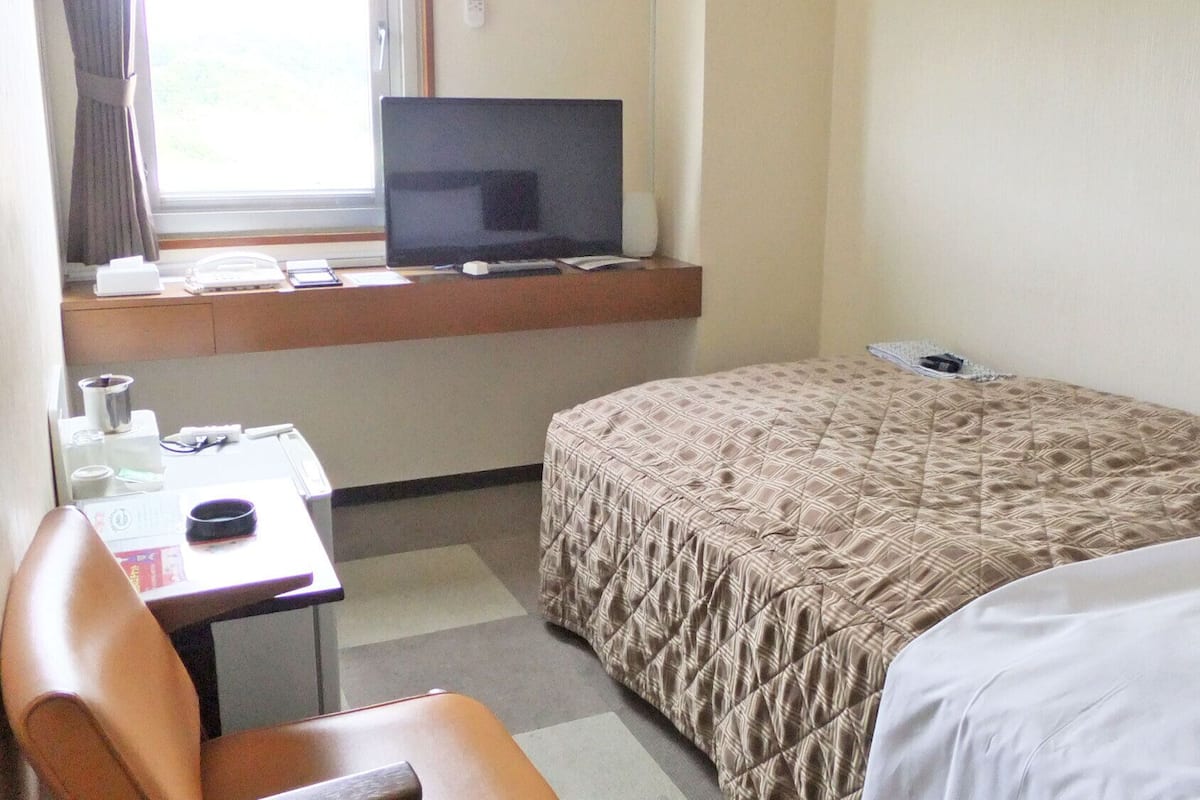 A hotel room with a bed, a desk with a computer, a chair, and a window with curtains.