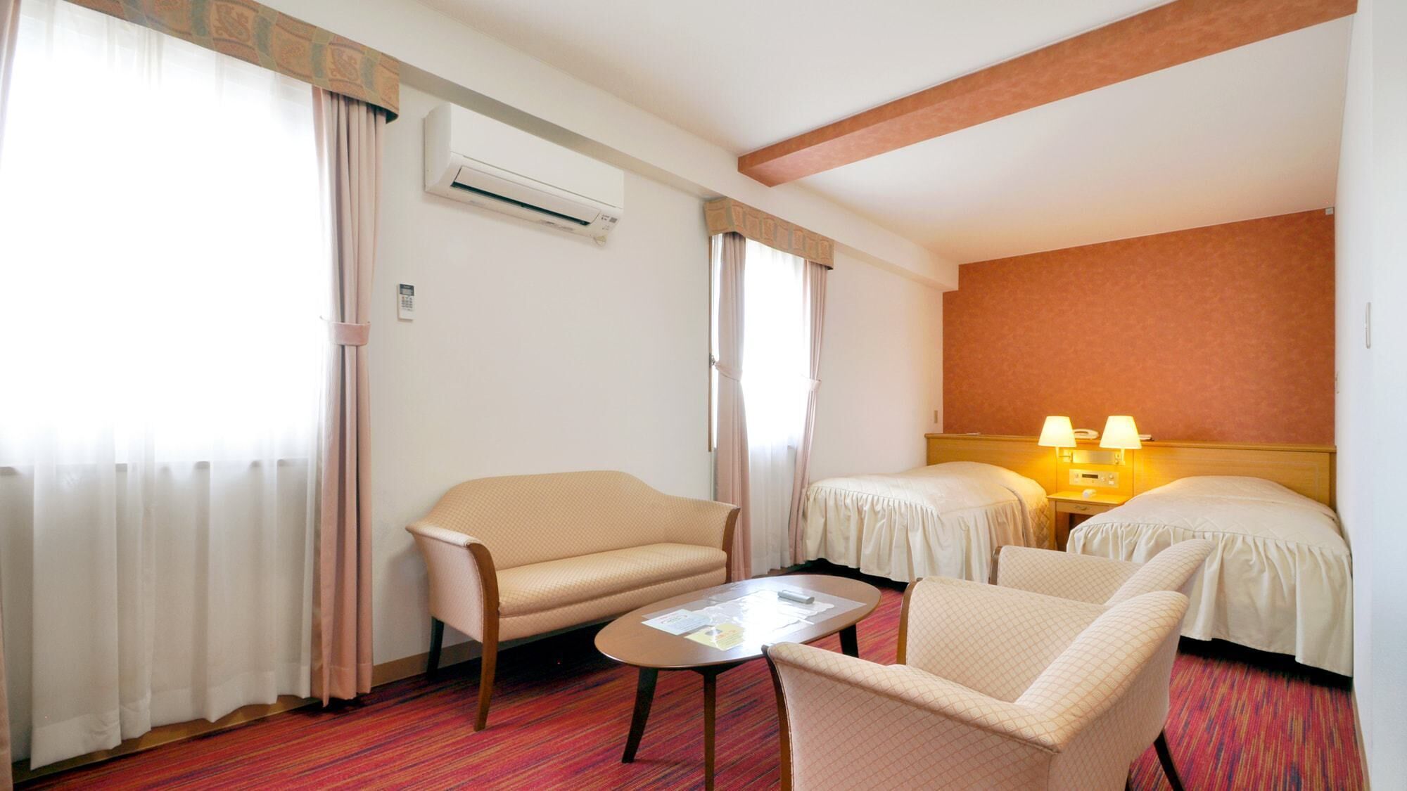 A hotel room with a sofa, two single beds, a small table, and an air conditioning unit.