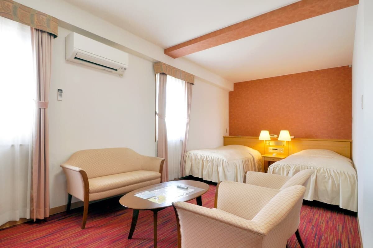 A hotel room with a sofa, two single beds, a small table, and an air conditioning unit.