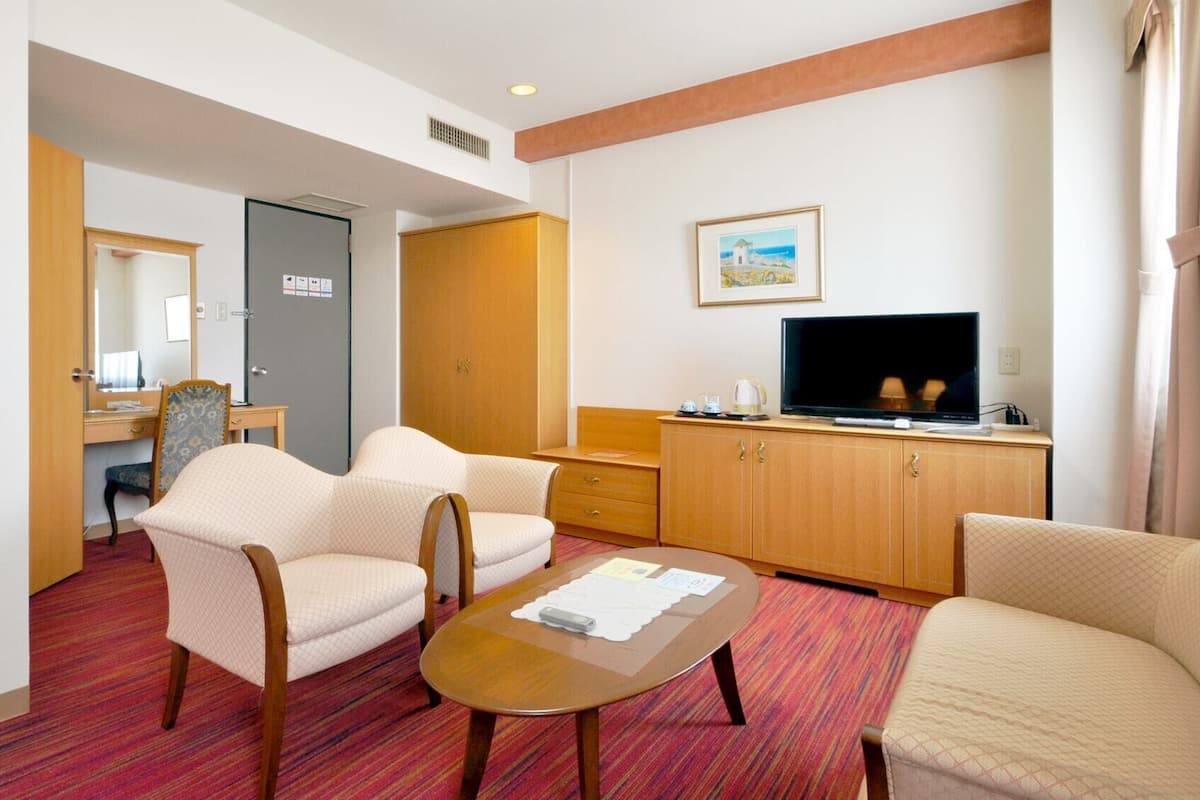 A hotel room with a bed, two armchairs, a small table, a TV, a desk with a chair, and a door.