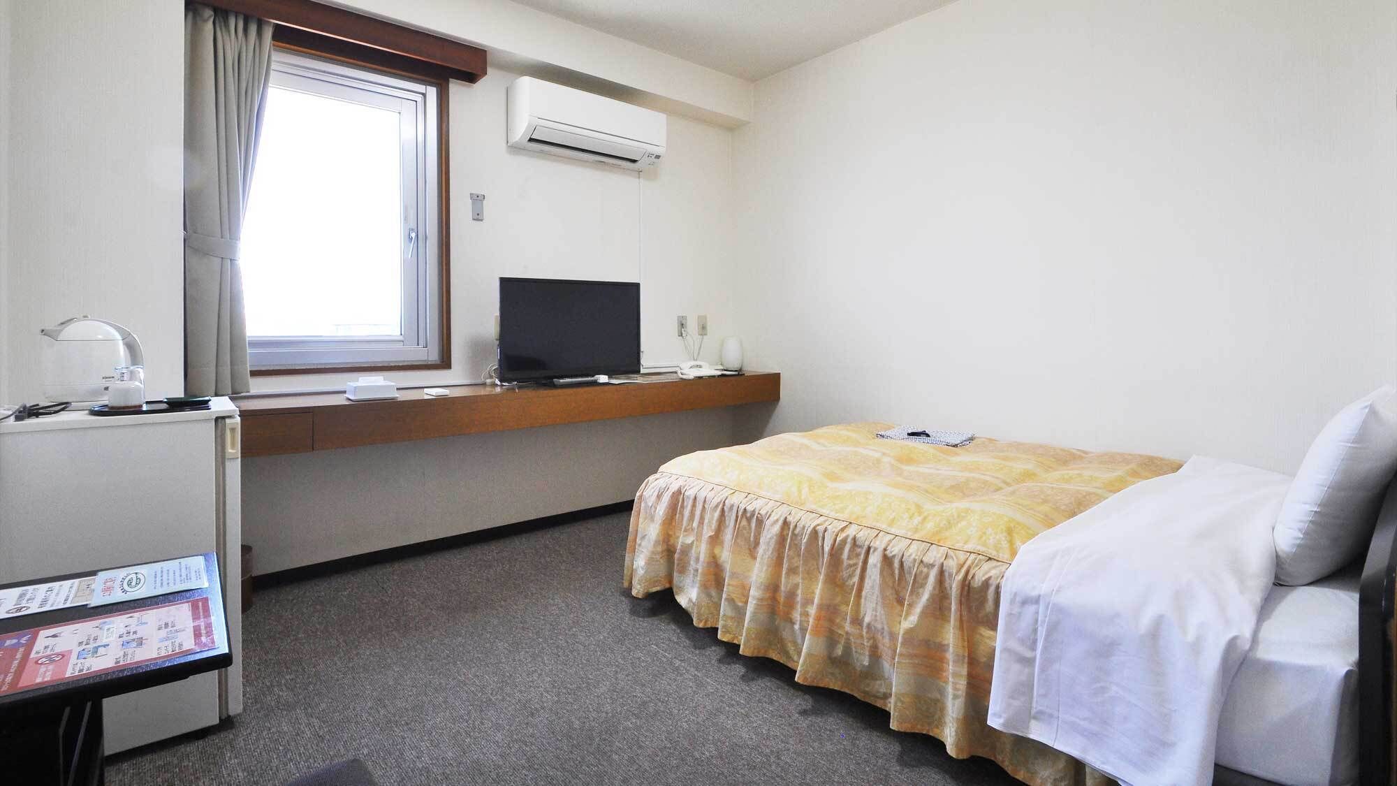 A hotel room with a bed, a desk with a computer, a window with curtains, and an air conditioning unit.
