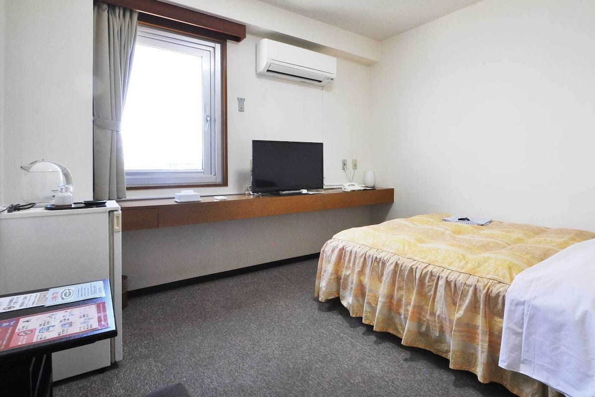 A hotel room with a bed, a desk with a computer, a window with curtains, and an air conditioning unit.