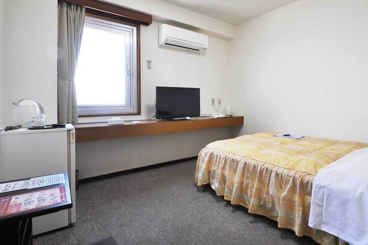 A hotel room with a bed, a desk with a computer, a window with curtains, and an air conditioning unit.