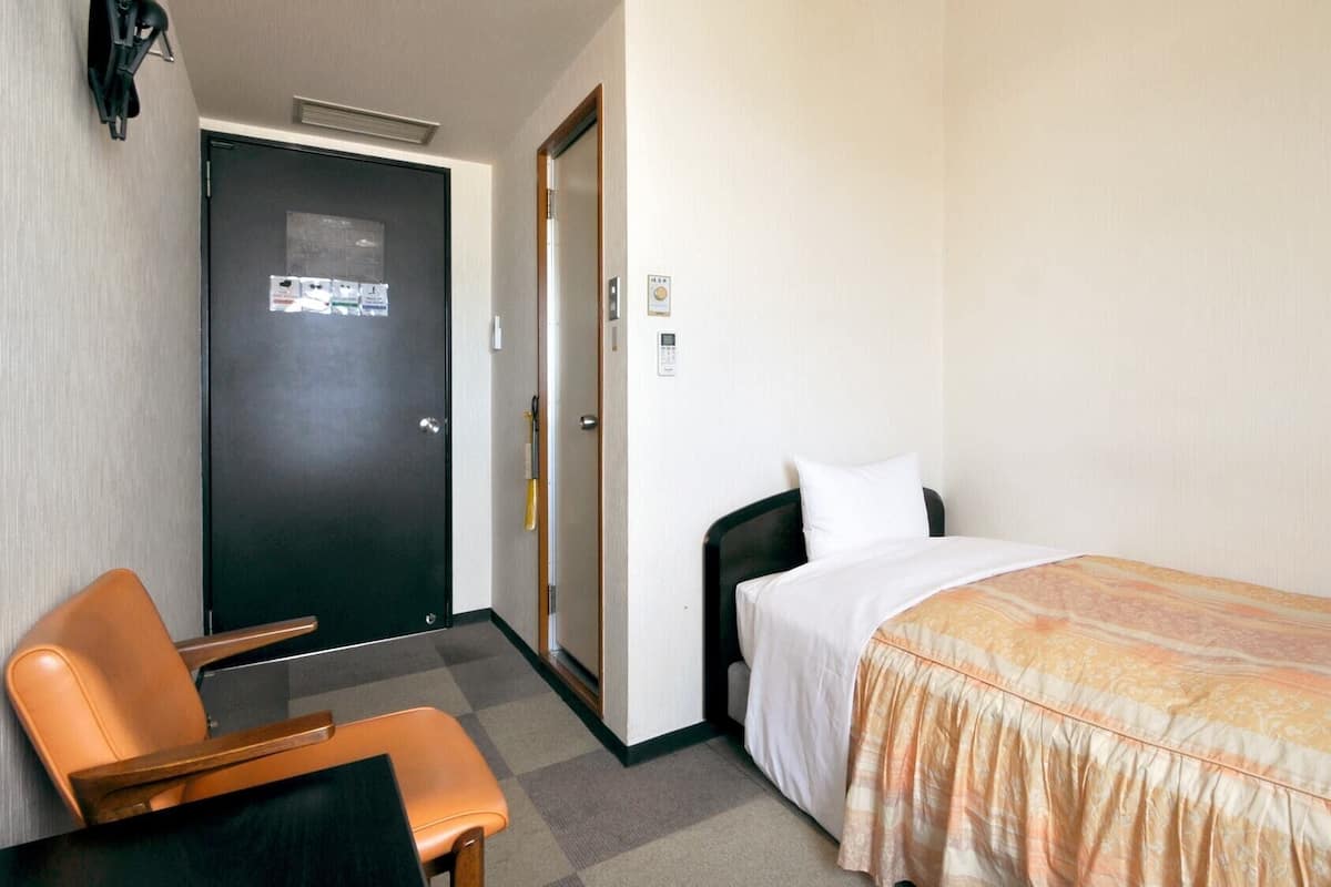 A hotel room with a bed, a chair, a desk, a door, and a wall-mounted phone.
