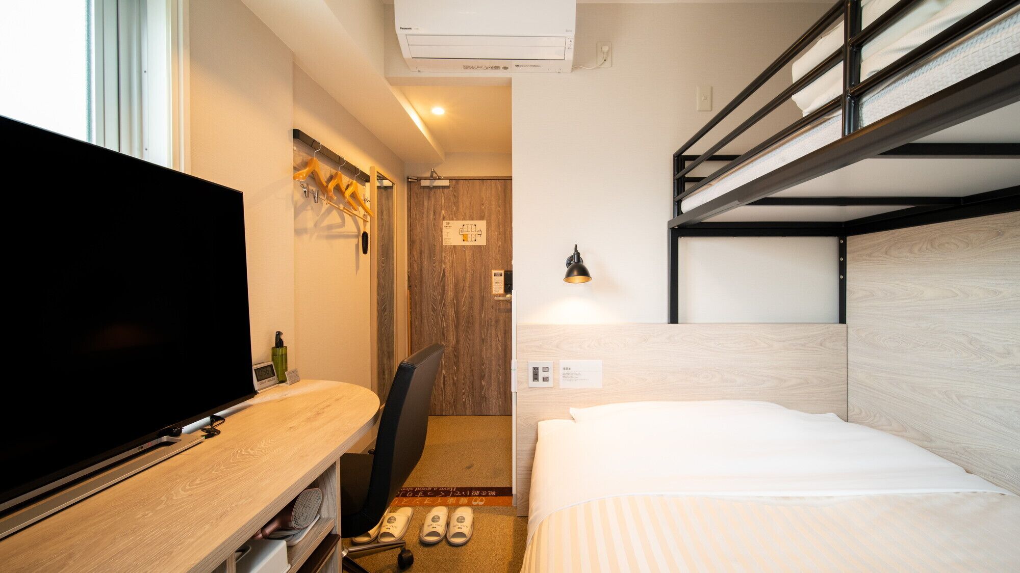 A compact hotel room with a bunk bed, a desk with a flat-screen TV, a chair, a small rug, and a wall-mounted lamp.