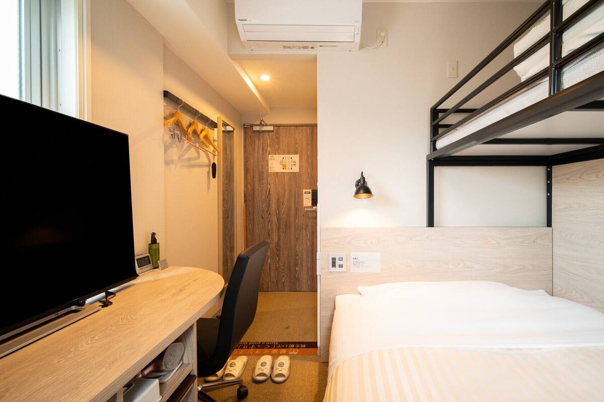 Een compacte hotelkamer met een stapelbed, een bureau, een flatscreen-televisie, een stoel, een klein vloerkleed en een wandlamp.