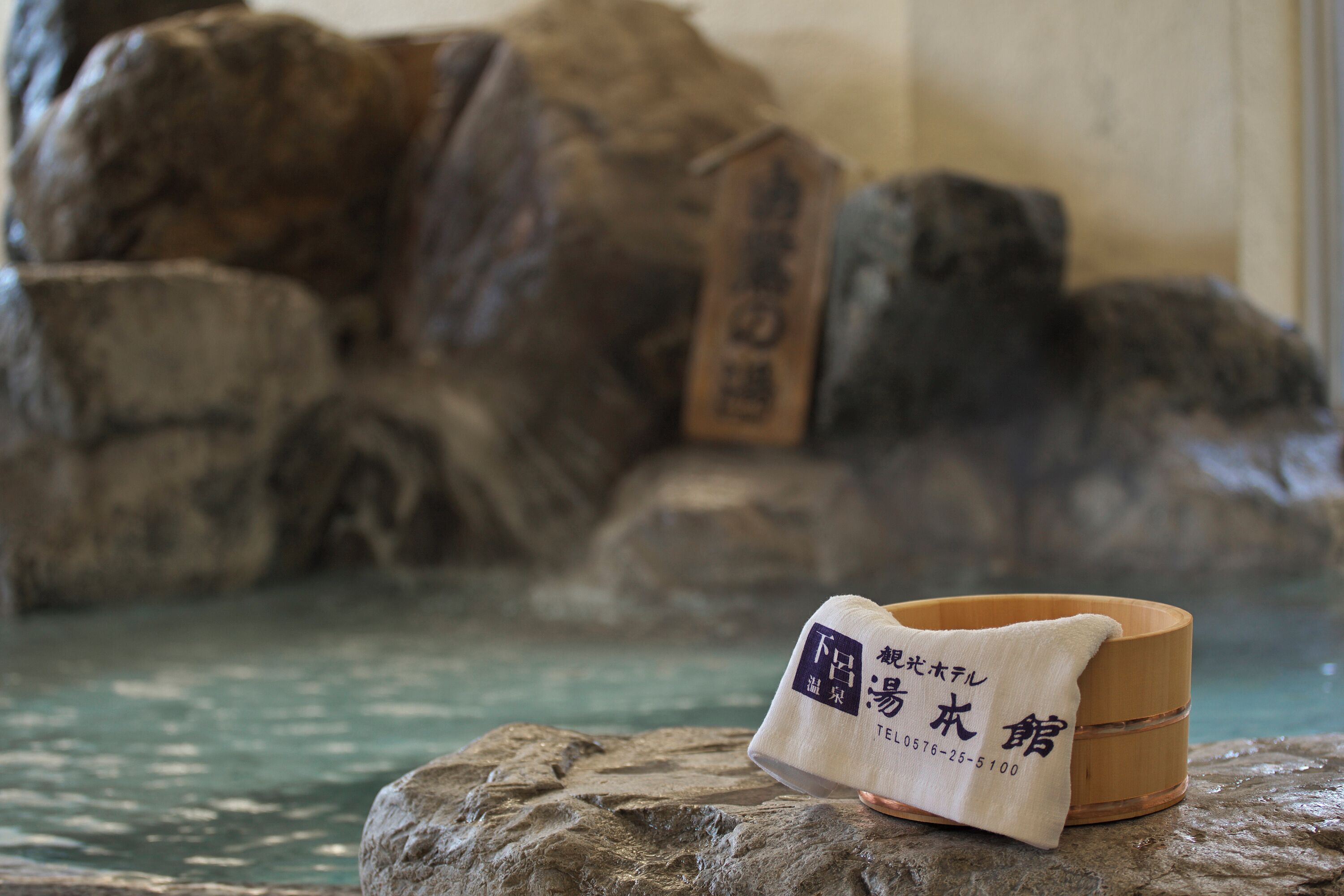 Un cuenco de piedra con una toalla y un recipiente de madera, probablemente para agua caliente, en un entorno natural entre rocas y agua.