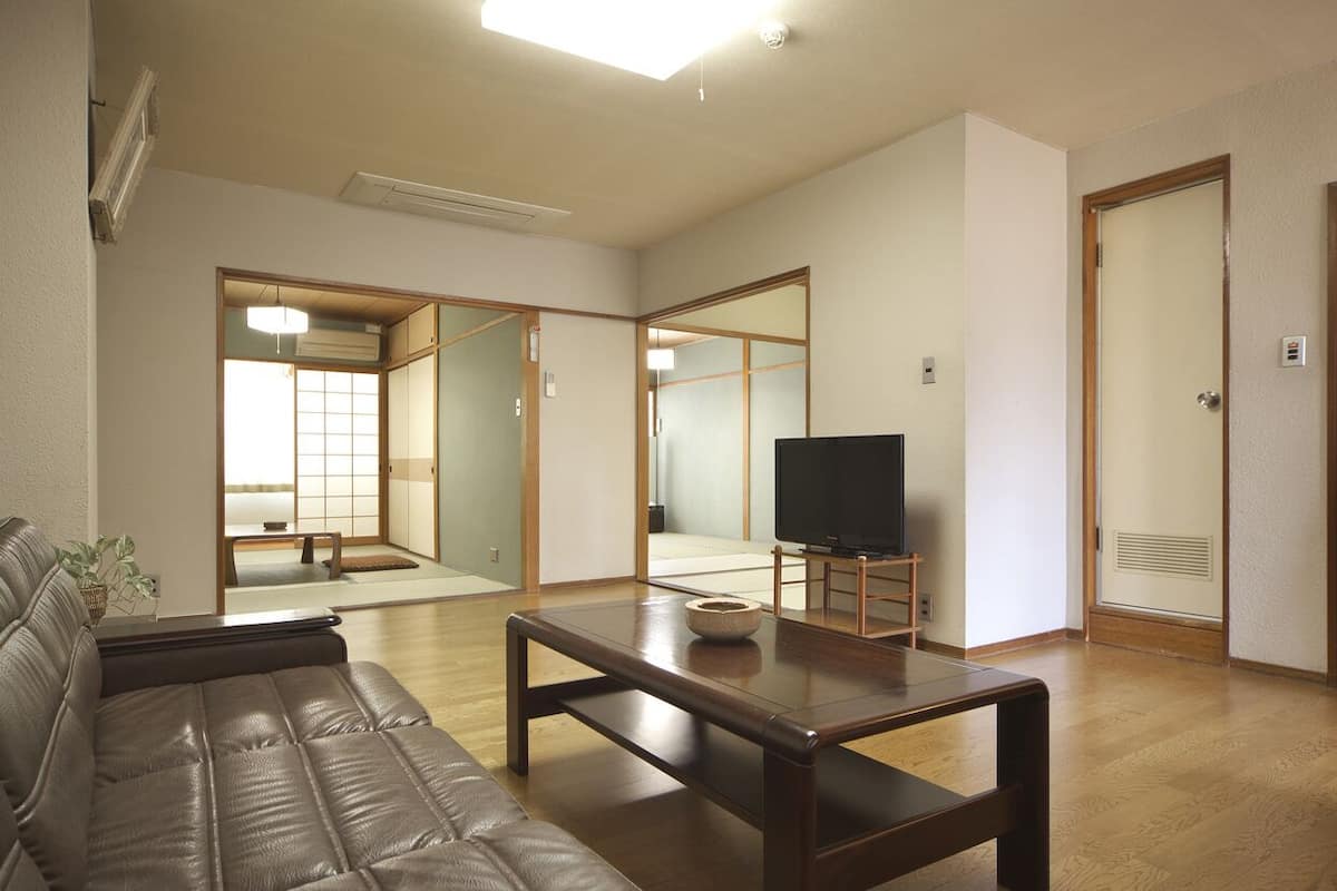 Una sala de estar con sofá, una mesita de centro, televisor y un puerta corredera que da a una habitación japonesa tradicional.