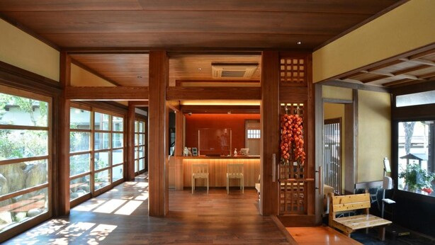 A spacious hallway with wooden flooring and large windows allowing natural light. A reception desk is visible in the distance, and there is a seating area with a bench on the right. - Yoshino Ryokan (Hitoyoshi)