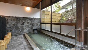 A traditional Japanese onsen with a stone wall, a wooden ceiling, and a view of a bamboo fence outside. - Yoshino Ryokan (Hitoyoshi)