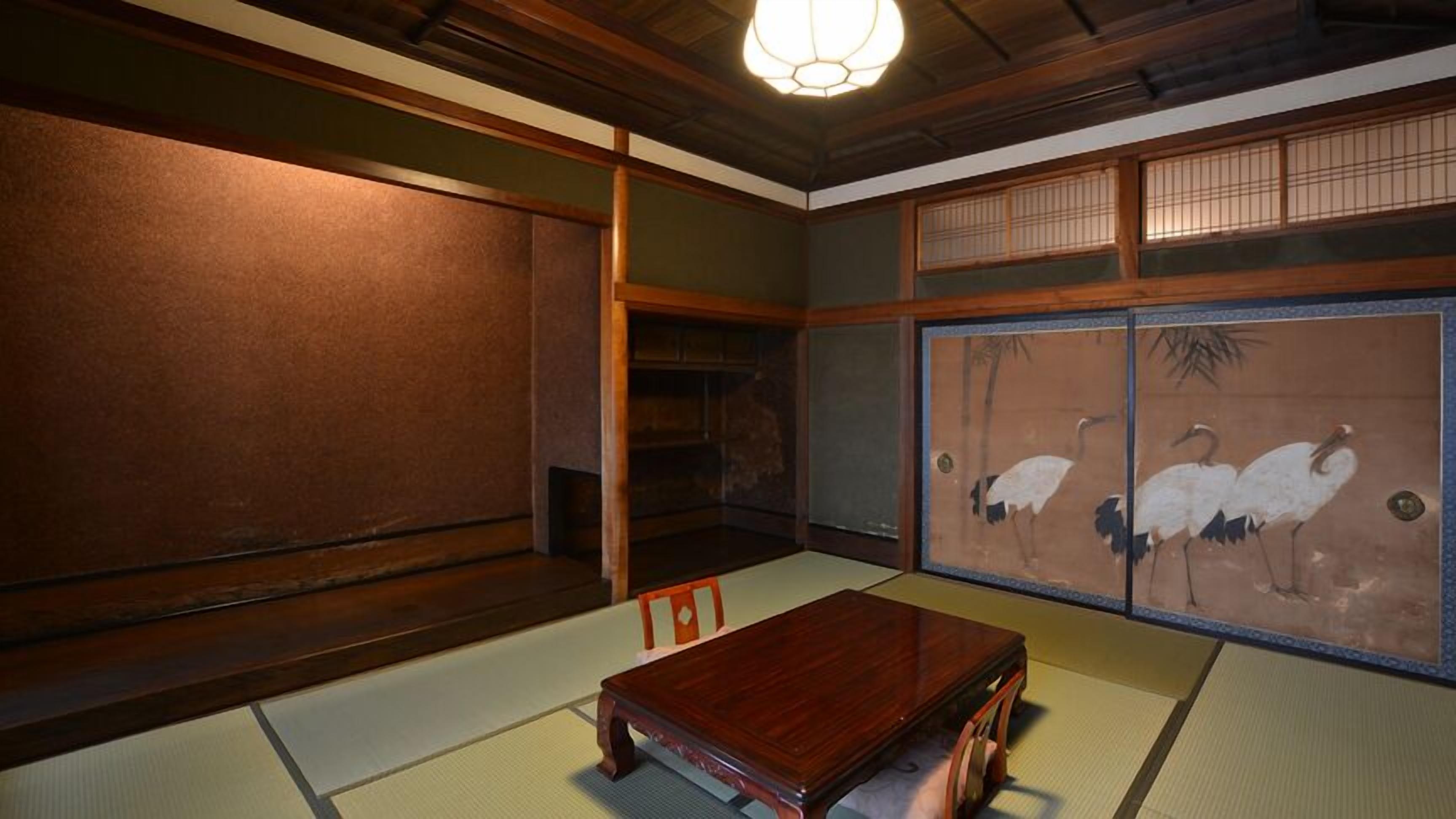 Une pièce de style japonais traditionnel, avec un sol recouvert de tatamis, une basse table en bois, des portes coulissantes ornées d’un motif naturel et un luminaire suspendu au plafond.