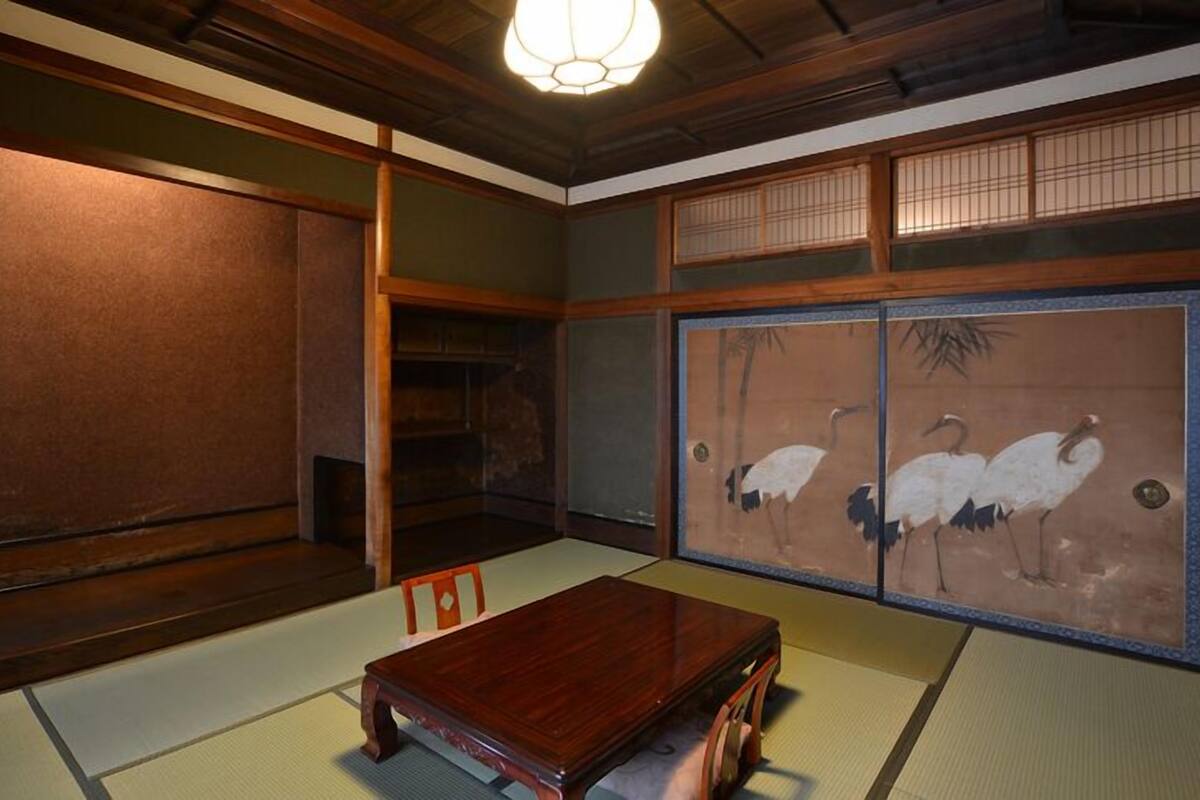 Ein traditioneller japanischer Raum mit Tatami-Boden, einem niedrigen Holztisch, Schiebetüren mit einer Naturdarstellung und einer Deckenleuchte.