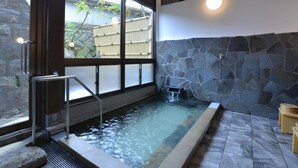 A spa area with a stone wall, a hot tub, and a window with a view of a bamboo fence. - Yoshino Ryokan (Hitoyoshi)