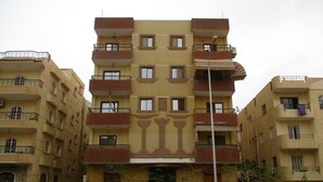 Un immeuble résidentiel à plusieurs étages, doté de balcons, d’une entrée principale et d’un espace de stationnement avec des voitures.
