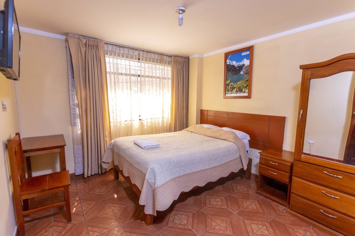 A bedroom with a bed, wooden dresser, bedside table, and a television mounted on the wall.