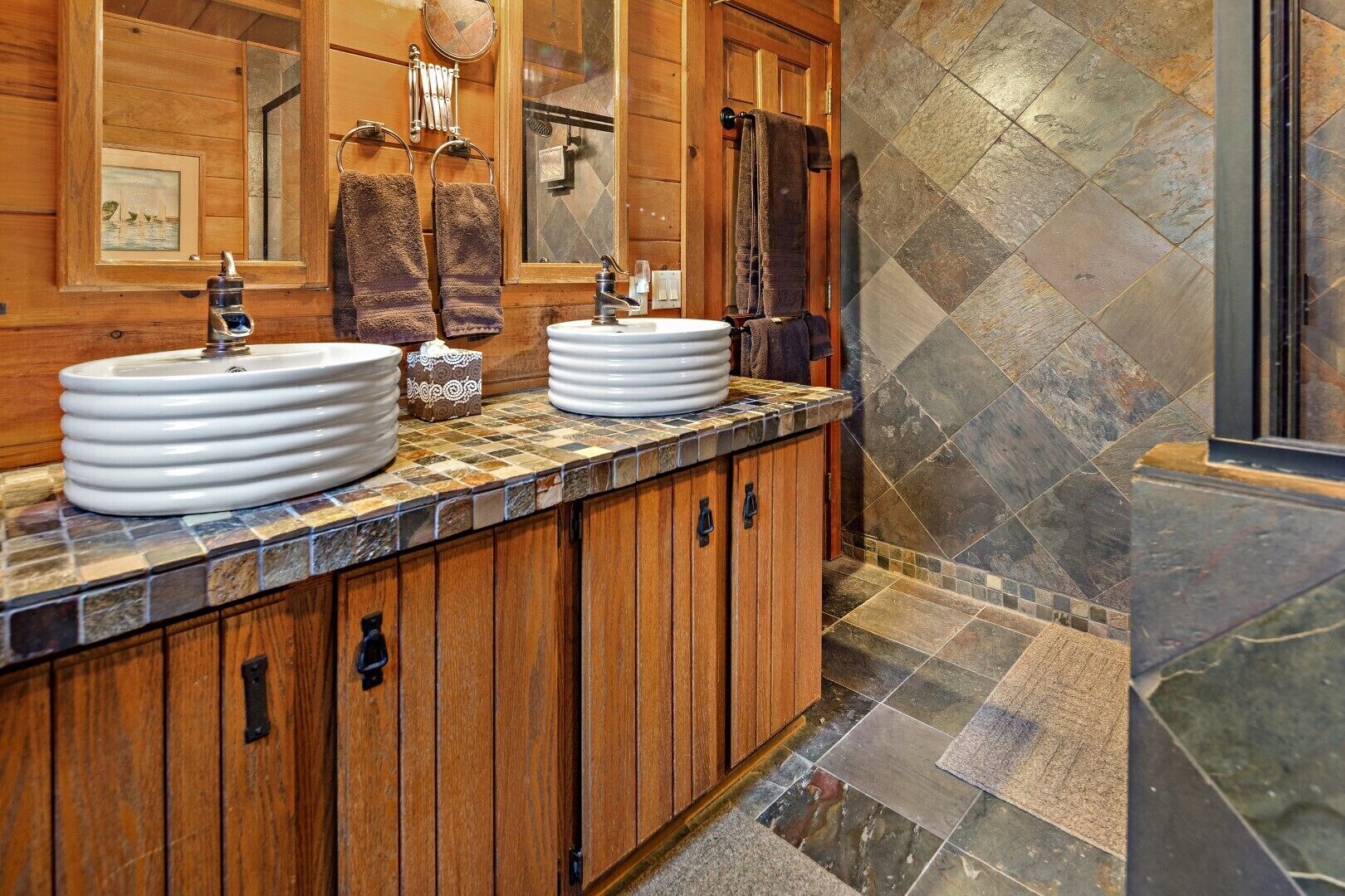Un baño con armarios de madera, dos lavamanos blancos tipo recipiente y piso de baldosas de piedra.