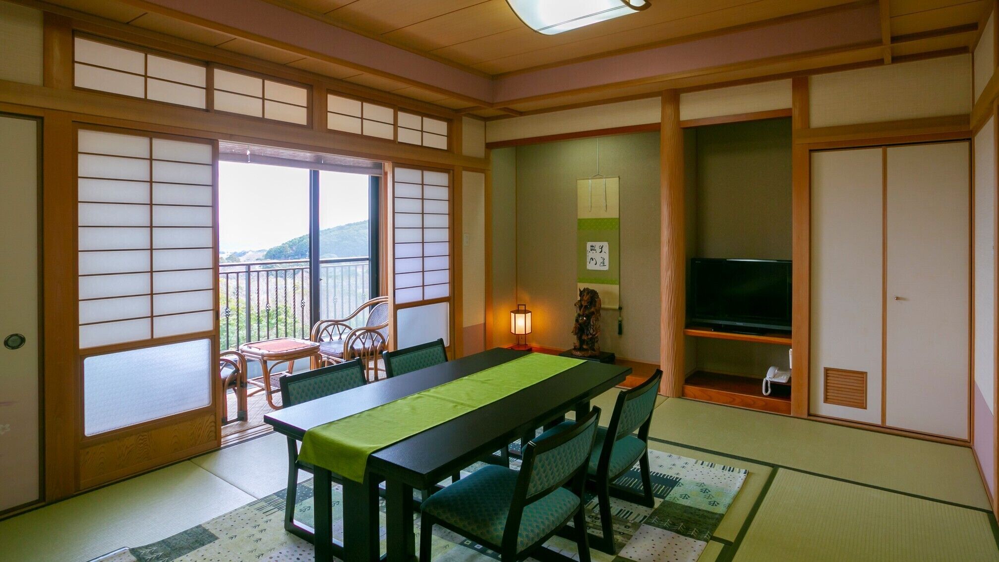 Une pièce de style japonais traditionnel, avec un sol recouvert de tatamis, une petite table recouverte d’une natte verte, des chaises en bois, un téléviseur et une porte coulissante donnant sur l’extérieur.