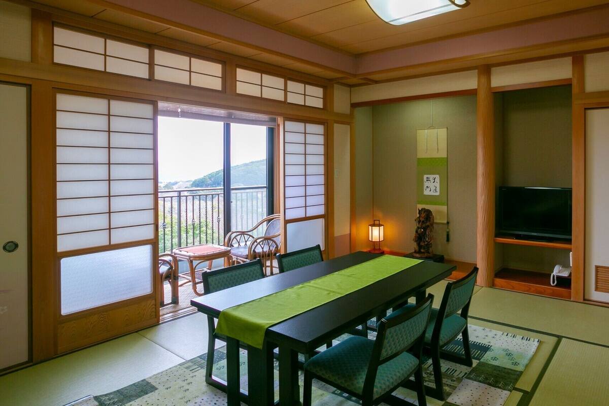 A traditional Japanese room with tatami flooring, a low table with a green mat, wooden chairs, a television, and a sliding door with a view of the outdoors.