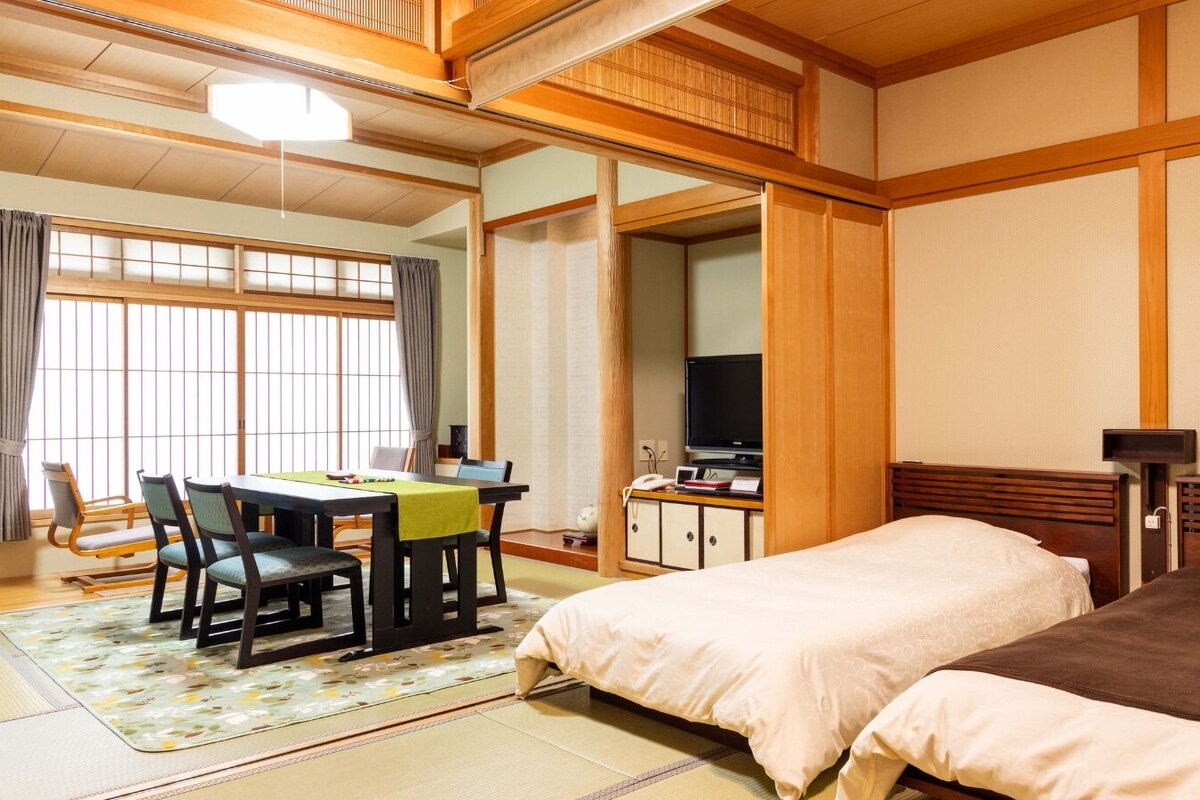 A traditional Japanese-style hotel room with tatami flooring, a futon bed, a desk with a television, and a sliding door.
