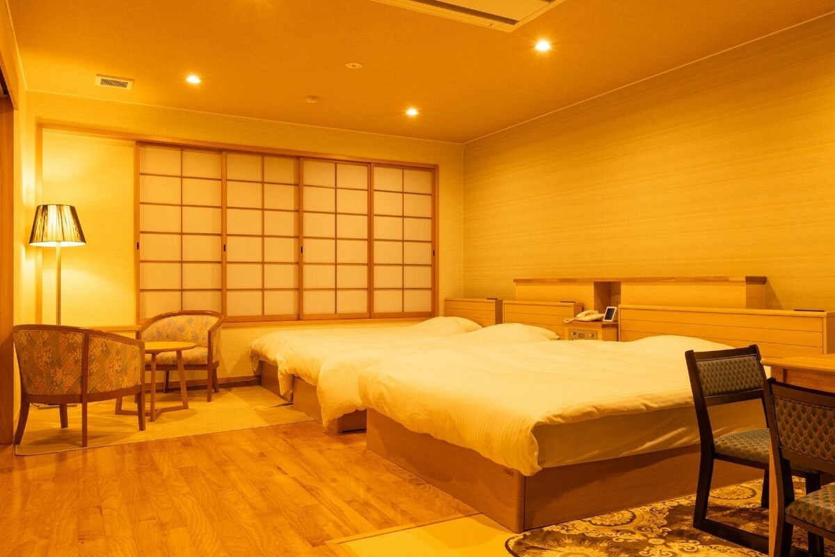A hotel room with two beds, a desk, chairs, a lamp, and a sliding door.