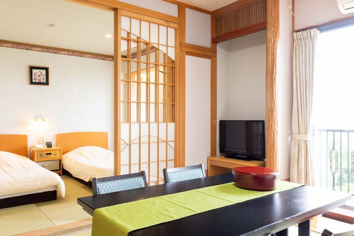 A hotel room with two beds, a television, a dining table, and a sliding door.
