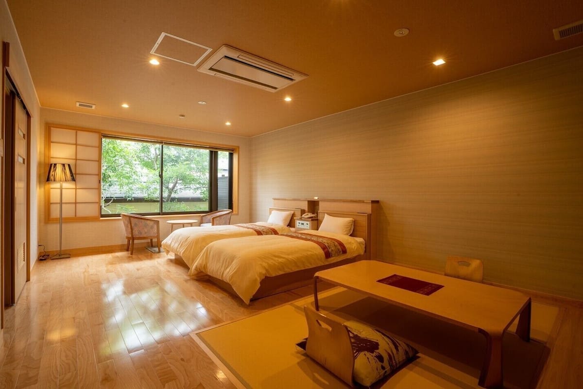 A hotel room with two beds, a desk, a chair, a bedside table, and a window with a view of greenery.