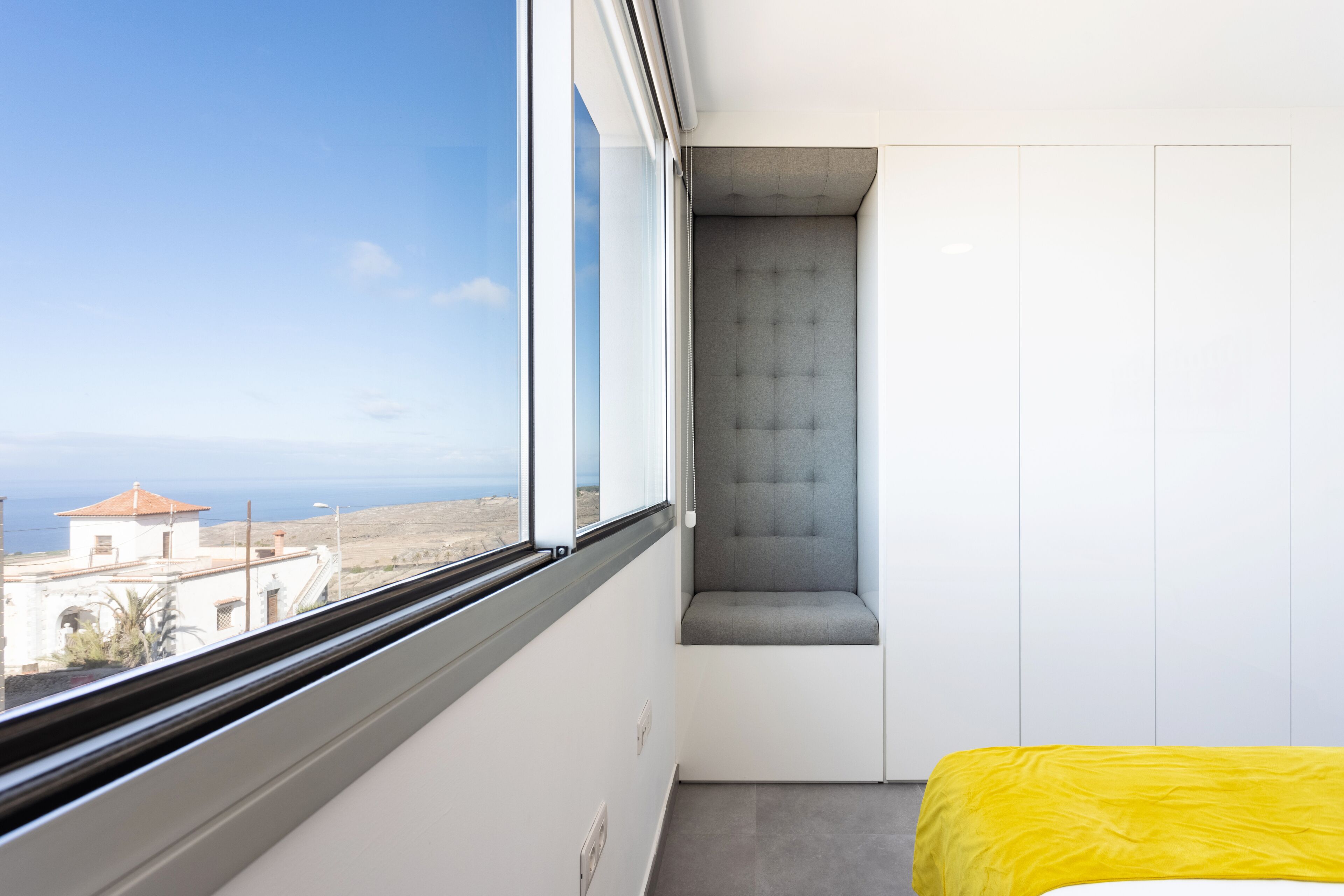 A room with a window, a bed with a yellow bedspread, and a grey upholstered bench.