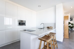 Una cocina moderna con armarios blancos, horno empotrado, pileta de acero inoxidable y encimera de mármol. Hay tres taburetes de madera en la isla.
