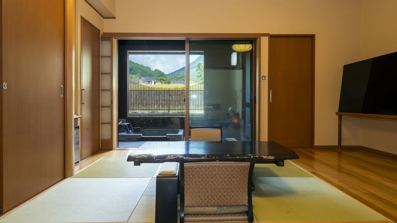 Habitación con suelo de tatami, puerta corredera a un balcón, una mesa negra con una silla y un televisor en la pared.
