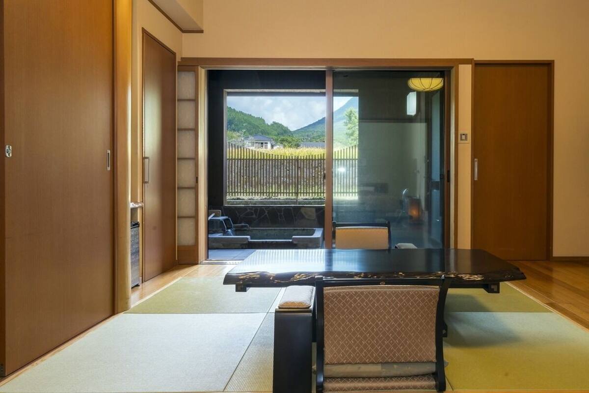 A room with tatami flooring, a sliding door leading to a balcony, a black table with a chair, and a TV on the wall.