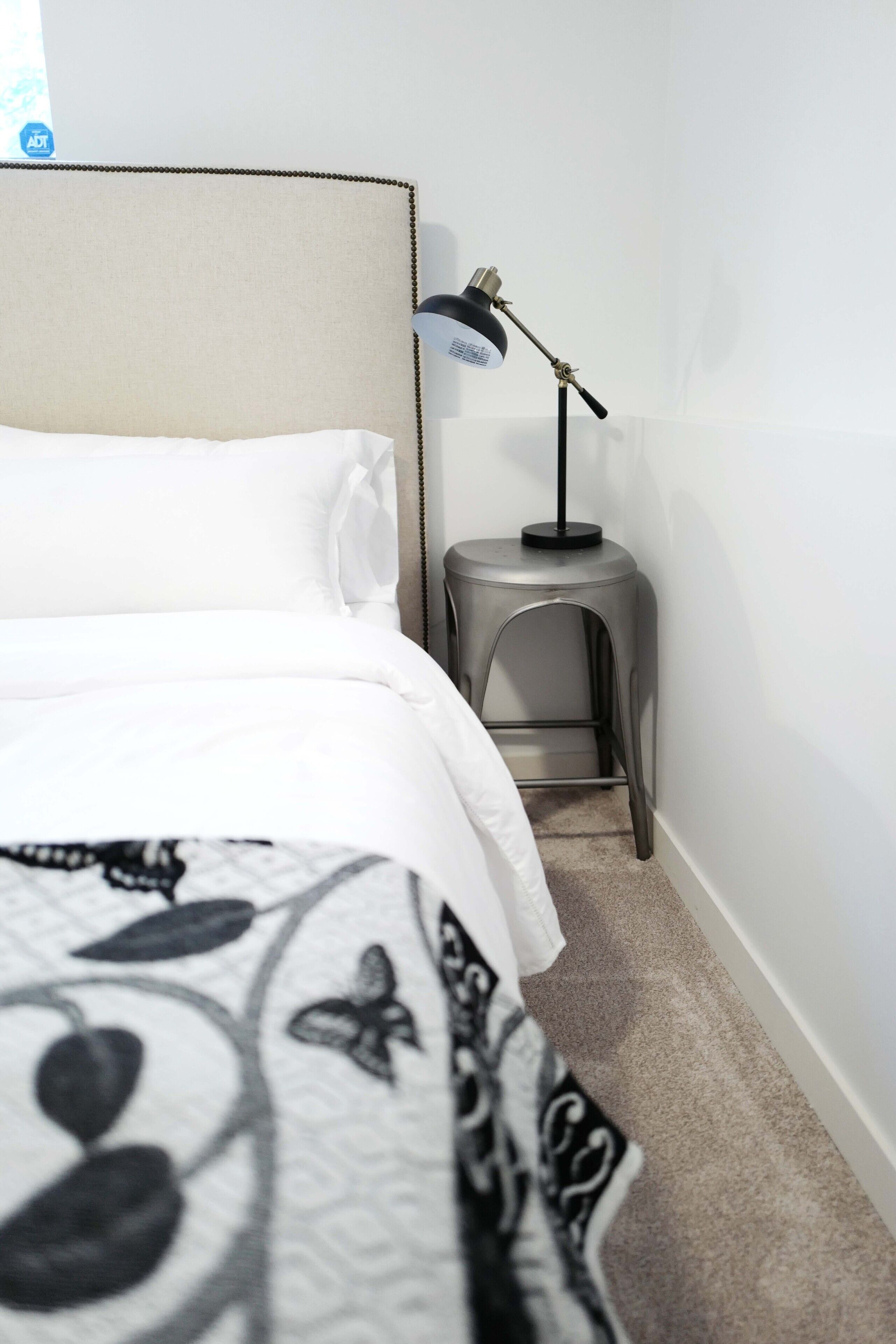 Un lit bien fait, avec une literie blanche et une couverture à motifs noir et blanc. Il y a un petit tabouret avec une lampe à côté du lit.