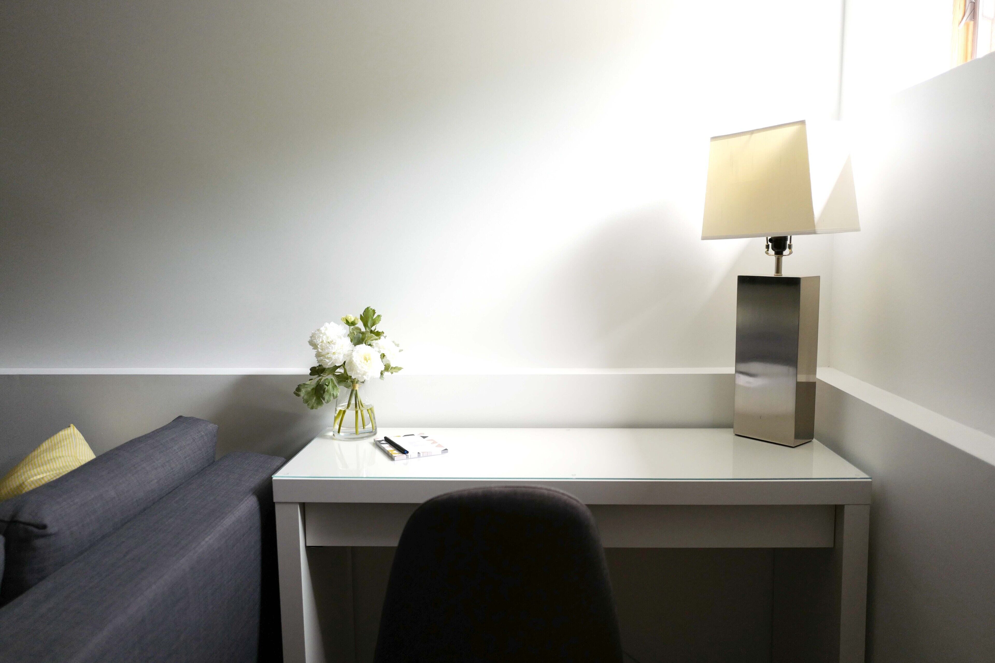 Une chambre moderne avec un bureau blanc, une chaise noire, un petit vase avec des fleurs blanches et une lampe.