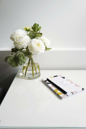 Un florero de vidrio transparente con flores blancas y hojas verdes sobre una mesa blanca.