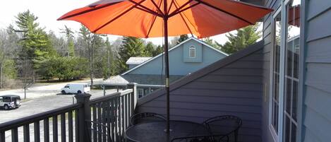 Un balcon avec une table et des chaises en métal, un parasol rouge et une vue sur une rue et des maisons.