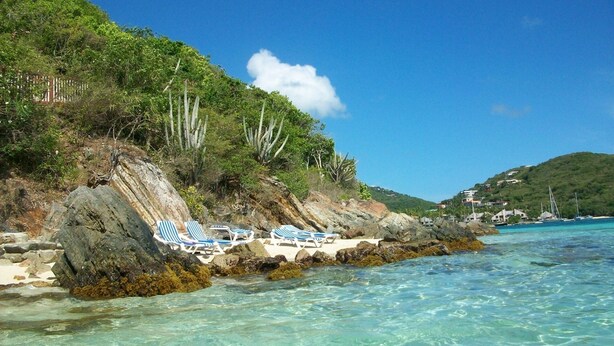 Una playa con aguas turquesas cristalinas, afloramientos rocosos y algunas sillas de playa en la orilla.