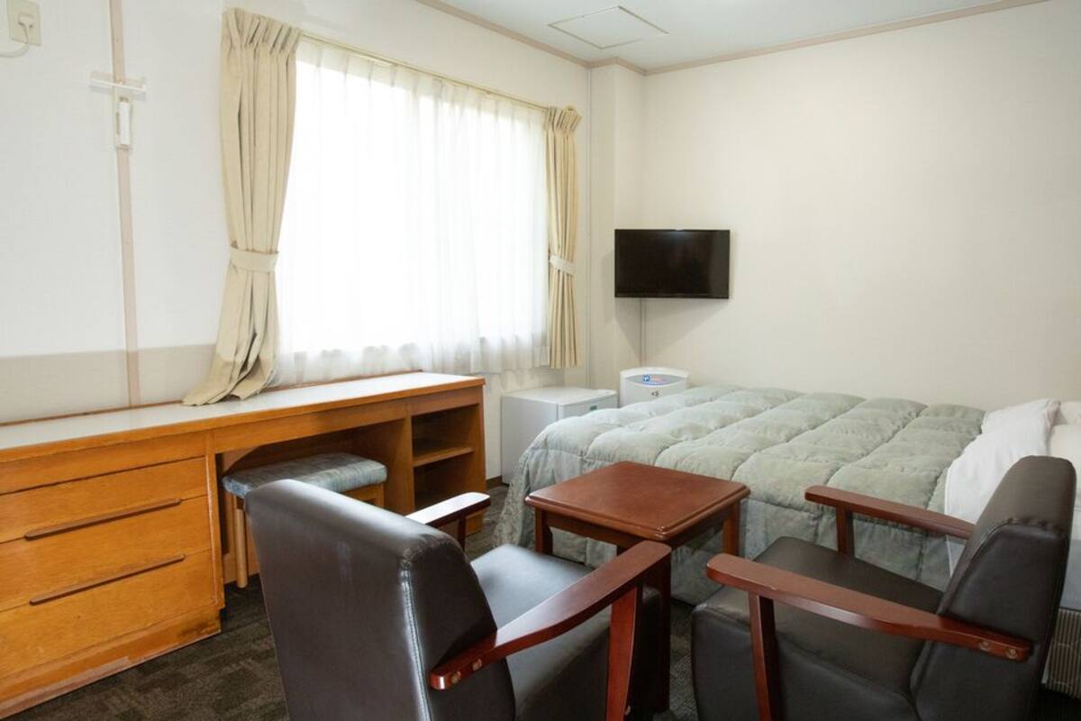 Habitación de hotel con cama, escritorio, silla, televisor y ventana con cortinas.
