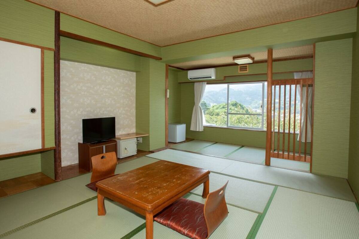 Una habitación tradicional japonesa con suelo de tatami, una mesa baja con cojines, un televisor y una puerta corredera con vistas a la vegetación.