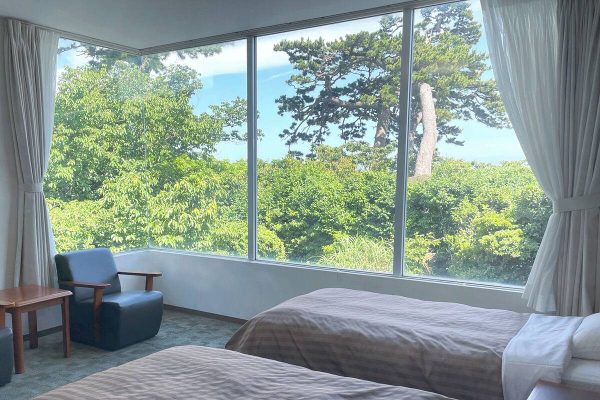 Habitación de hotel con un ventanal grande, dos camas, una silla, una mesita y vistas a zonas verdes.