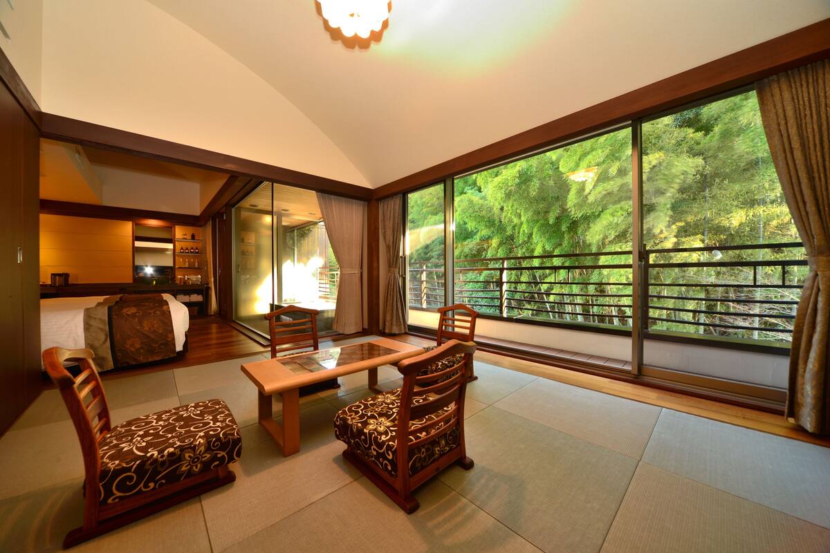 A spacious room with a large sliding glass door leading to a balcony, a bed, a dining table with chairs, and a small sofa.