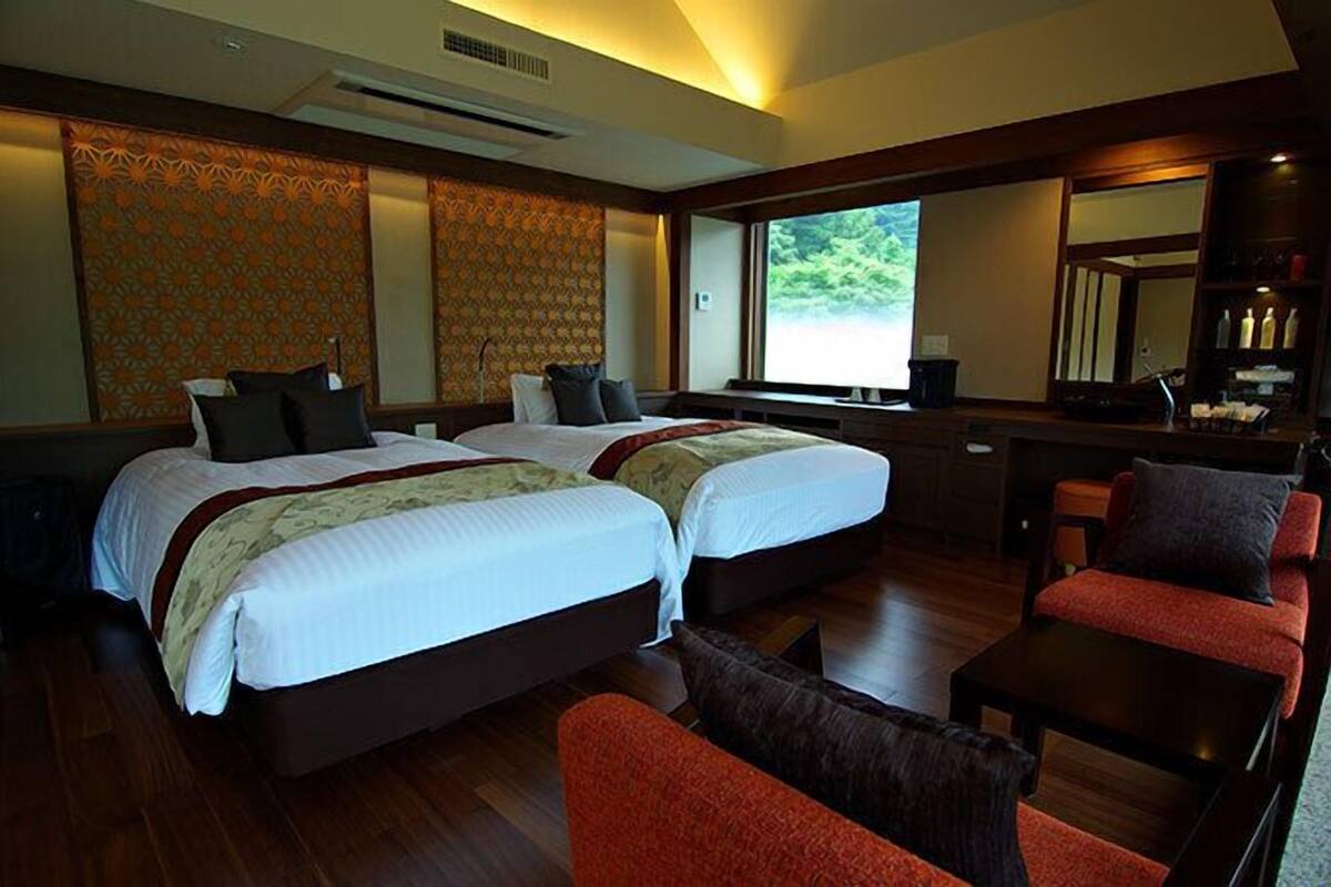 A hotel room with two beds, a desk, a chair, and a large window offering a view of greenery.