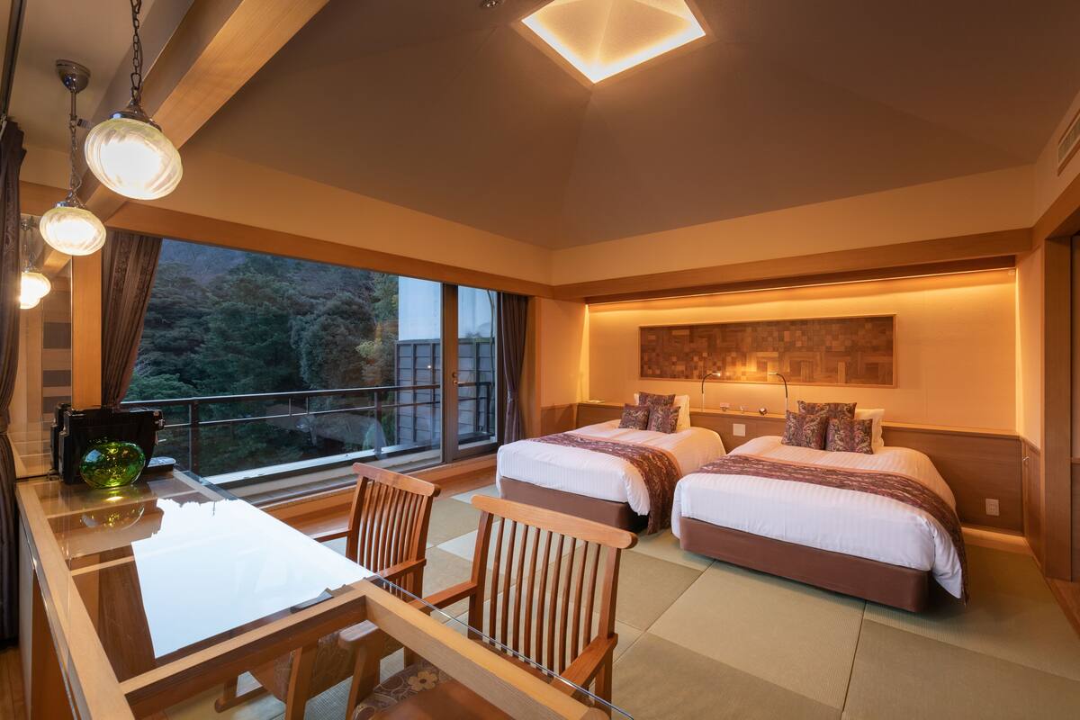 A modern hotel room with two beds, a dining area, and a large window with a view of trees.