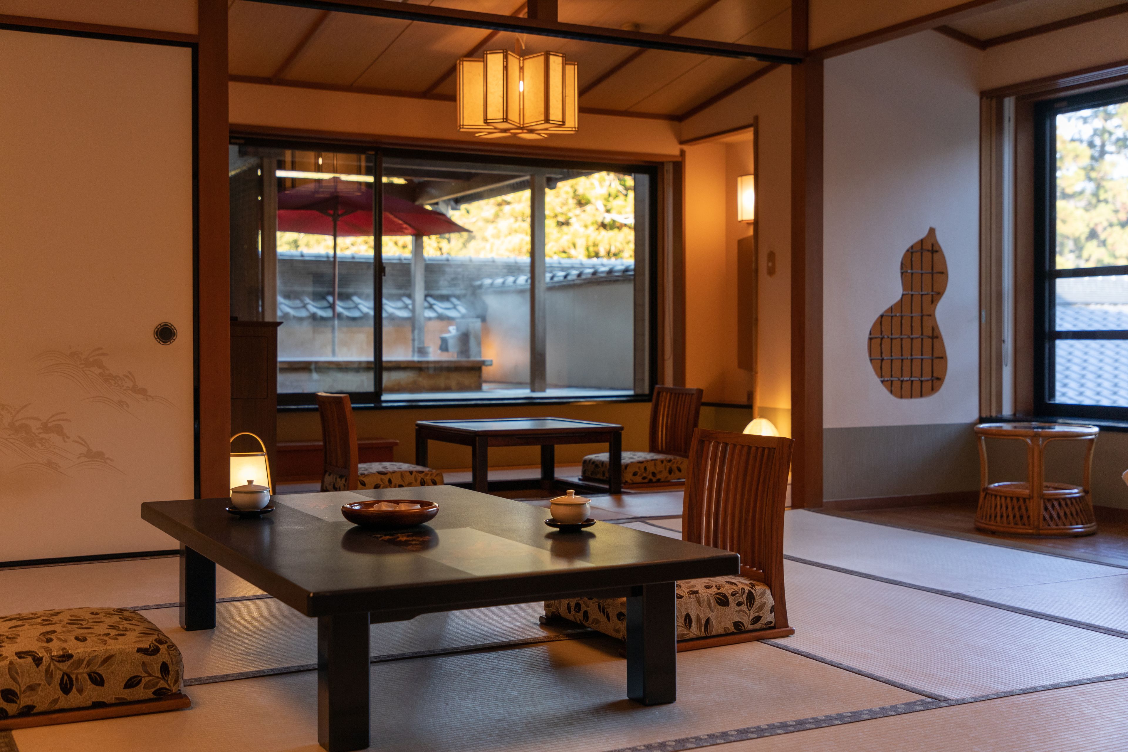 A traditional Japanese room with tatami flooring, a low table with a bowl and a candle, and a view of a traditional building outside.