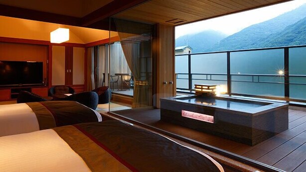 A hotel room with a large bed, a TV, a seating area with a table and chairs, and a balcony with a view of mountains and a lake. - Hakone Kamon (Hakone)